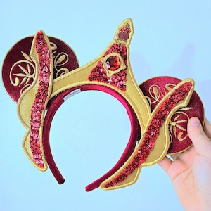 May include: A headband featuring two circular maroon velvet ears with gold embroidered patterns. A central gold crown-like structure sits between the ears, adorned with a red faceted gem and red sequins. The brand tag reads Antikapr.