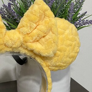 May include: Yellow fuzzy bow headband with a clear plastic headband. The headband is sitting on a white surface.