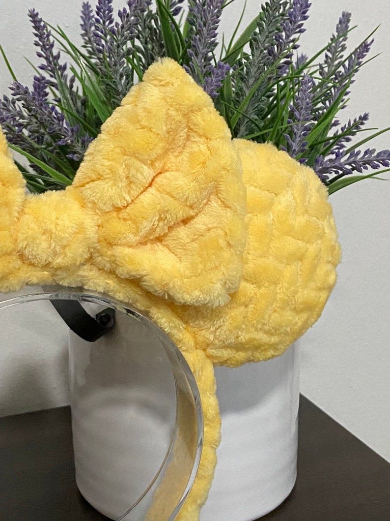 May include: Yellow fuzzy bow headband with a clear plastic headband. The headband is sitting on a white surface.