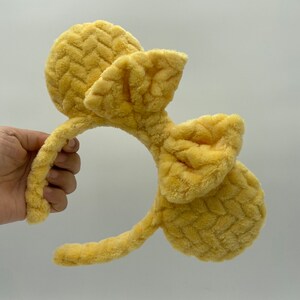 May include: Yellow plush headband with large, floppy ears and a bow. The headband is made of a soft, textured fabric.