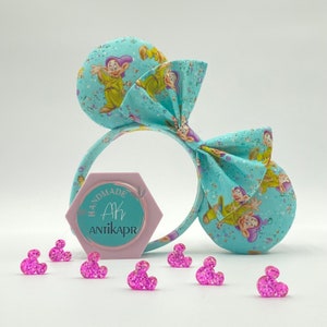 May include: A pair of blue and white fabric Minnie Mouse ears with a bow. The ears are decorated with a pattern of the seven dwarfs from the Disney movie Snow White and the Seven Dwarfs. The bow is tied in a large, floppy knot. The ears are handmade and have a pink label that says "Handmade Ak Antikapr".