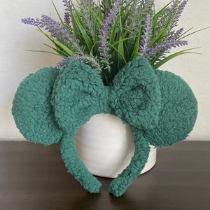 K&ouml;nnte beinhalten: Ein teal-farbenes Stirnband mit pl&uuml;schigen, abgerundeten Ohren und einer gro&szlig;en Schleife. Das Stirnband liegt vor einer wei&szlig;en Vase mit Lavendel und gr&uuml;nem Laub, was eine skurrile und verspielte &Auml;sthetik erzeugt.