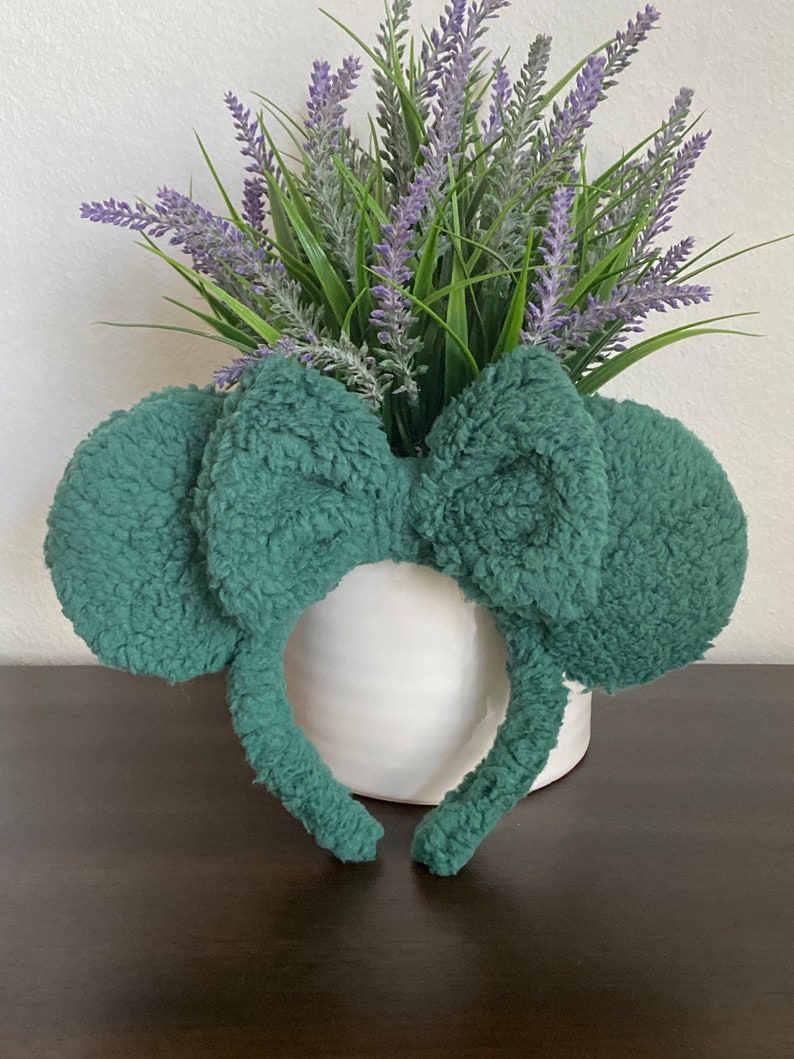 K&ouml;nnte beinhalten: Ein teal-farbenes Stirnband mit pl&uuml;schigen, abgerundeten Ohren und einer gro&szlig;en Schleife. Das Stirnband liegt vor einer wei&szlig;en Vase mit Lavendel und gr&uuml;nem Laub, was eine skurrile und verspielte &Auml;sthetik erzeugt.