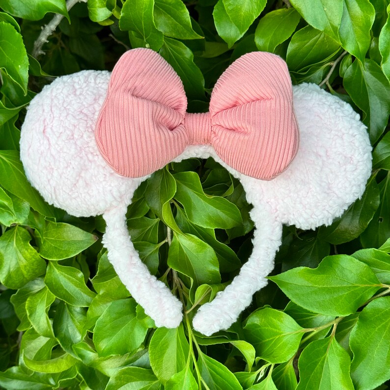 Pode incluir: Um par de orelhas de rato brancas e felpudas com um la&ccedil;o rosa. O la&ccedil;o &eacute; feito de um tecido canelado.