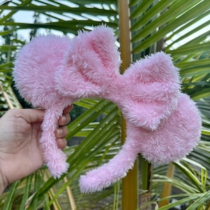 Op de afbeelding: Roze pluche Minnie Mouse oren haarband met een grote strik. De haarband is gemaakt van zacht, pluche materiaal.