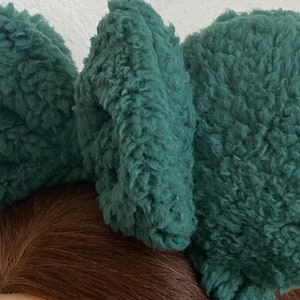 K&ouml;nnte beinhalten: Nahaufnahme eines dunkelblauen, flauschigen Stirnbandes mit drei gro&szlig;en, strukturierten, ohrenf&ouml;rmigen Akzenten. Das Stirnband besteht aus einem weichen, pl&uuml;schigen Material. Der Hintergrund ist hell.