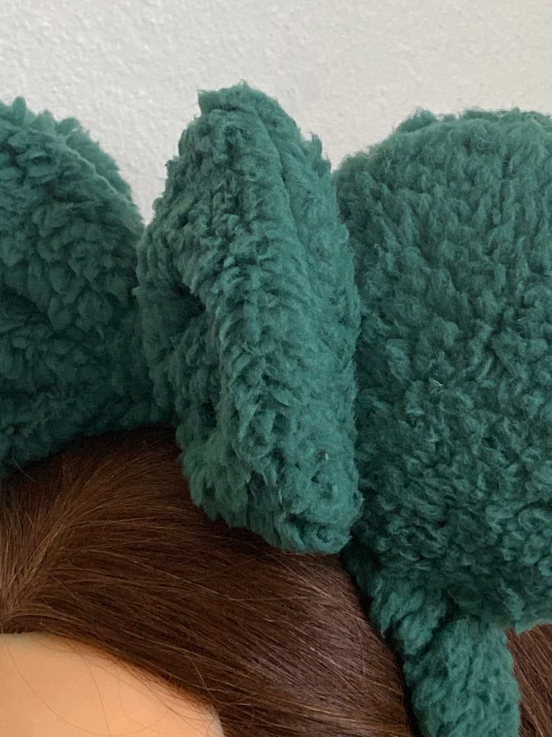 K&ouml;nnte beinhalten: Nahaufnahme eines dunkelblauen, flauschigen Stirnbandes mit drei gro&szlig;en, strukturierten, ohrenf&ouml;rmigen Akzenten. Das Stirnband besteht aus einem weichen, pl&uuml;schigen Material. Der Hintergrund ist hell.