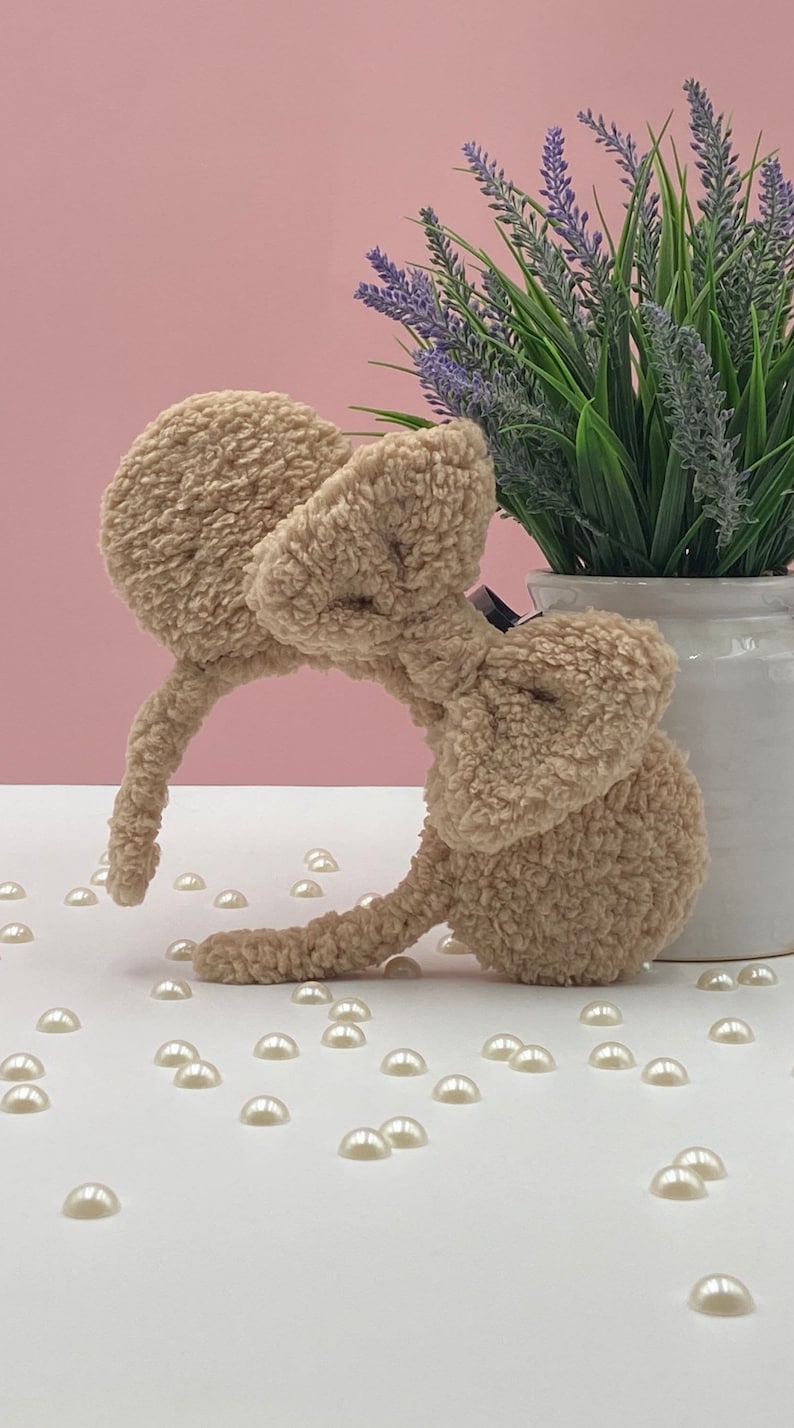 May include: A beige fuzzy headband with large bow and ears. The headband is on a white surface with white pearl-like beads scattered around it. A potted plant with purple flowers is in the background.