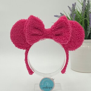 May include: Pink fluffy headband with a large bow. The headband is on a clear stand with a blue circle label that says "Handmade Ah AntikaPr".