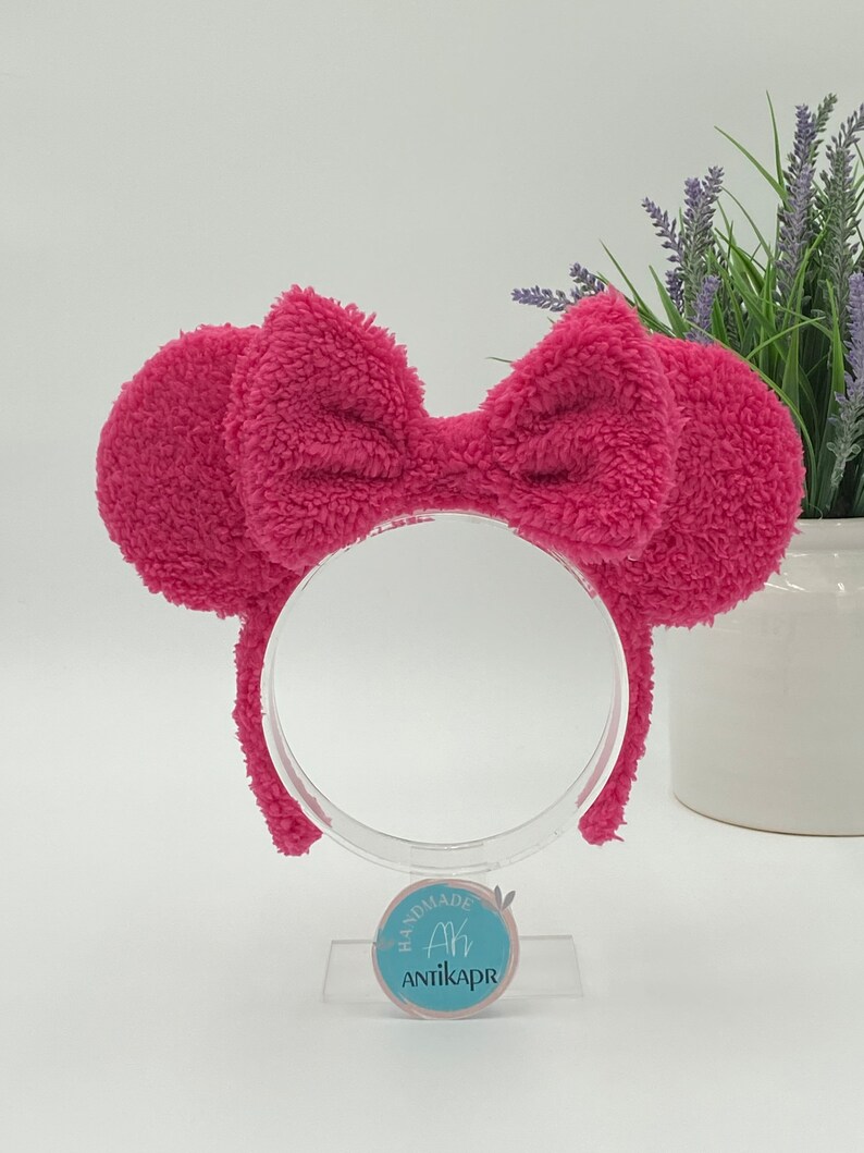 May include: Pink fluffy headband with a large bow. The headband is on a clear stand with a blue circle label that says "Handmade Ah AntikaPr".
