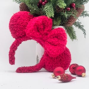 Puede incluir: Una diadema roja brillante y esponjosa con un gran lazo, que se asemeja a orejas de rat&oacute;n, se encuentra cerca de adornos navide&ntilde;os rojos y un arreglo festivo de pino y bayas. La diadema es un accesorio texturizado.
