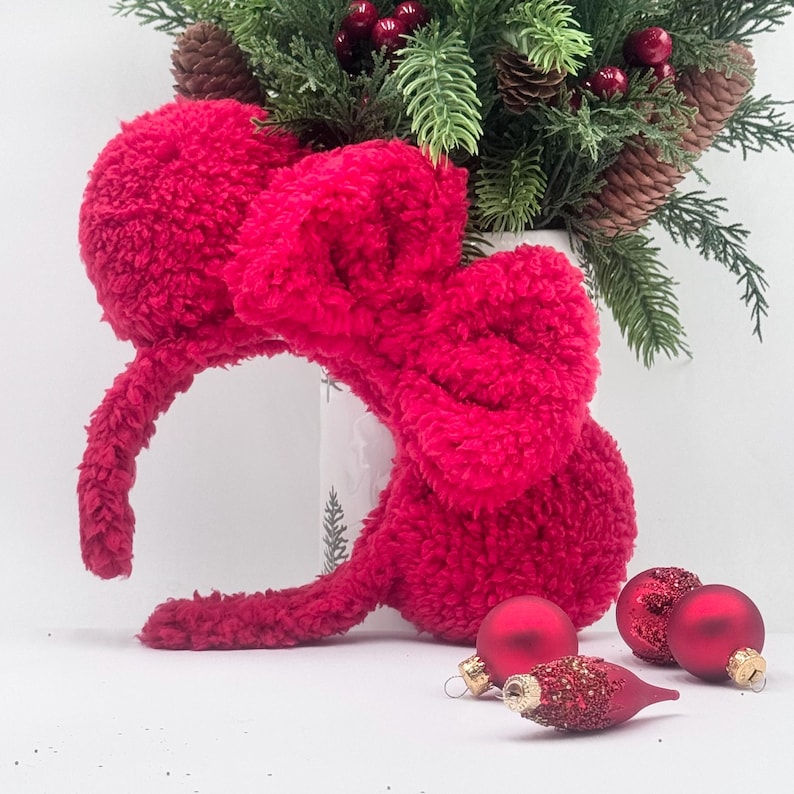 Puede incluir: Una diadema roja brillante y esponjosa con un gran lazo, que se asemeja a orejas de rat&oacute;n, se encuentra cerca de adornos navide&ntilde;os rojos y un arreglo festivo de pino y bayas. La diadema es un accesorio texturizado.