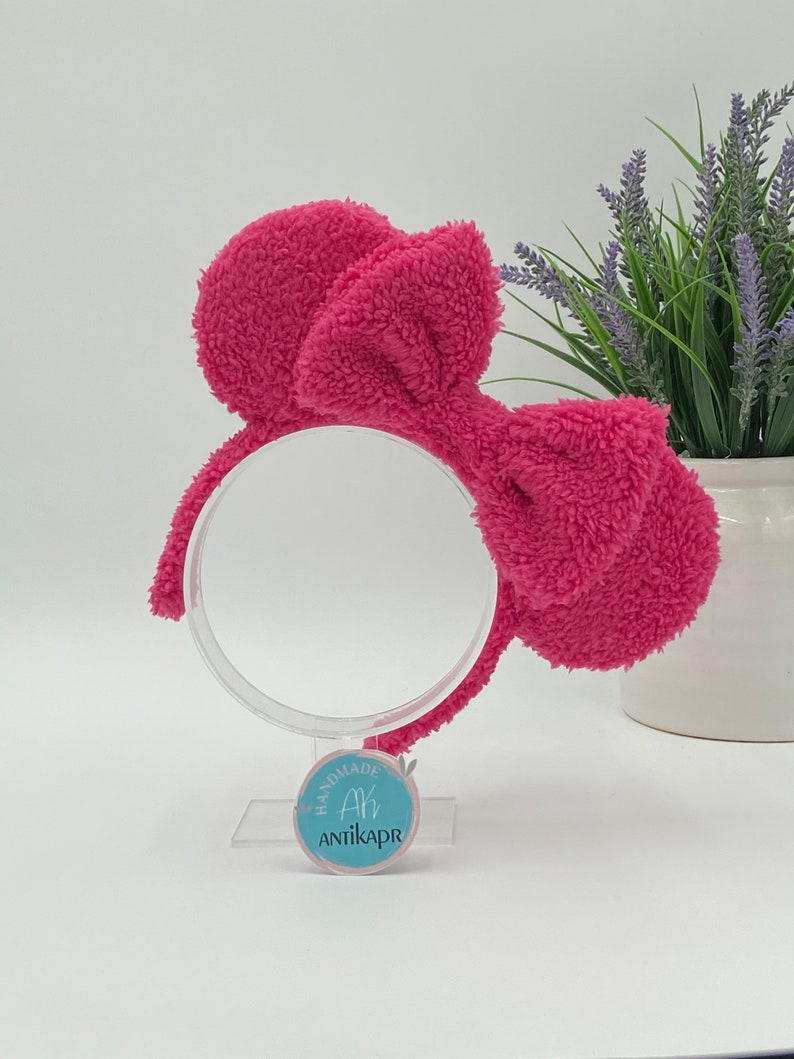 May include: A pink fluffy headband with a large bow. The headband is on a white stand with a blue circle label that says "Handmade Ah Antikapr".