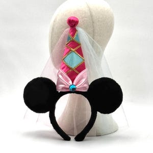 May include: A headband featuring two round black fuzzy ears and a central cone-shaped hat. The hat is pink and light blue with gold trim, a pink satin bow at the base, a blue gem, and a sheer white veil attached to the back of the cone structure.