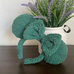 K&ouml;nnte beinhalten: T&uuml;rkisfarbenes Pl&uuml;sch-Mausohren-Stirnband. Die runden, flauschigen Ohren und das Stirnband sind t&uuml;rkisfarben. Das Stirnband liegt auf einer dunkelbraunen Oberfl&auml;che, mit einer wei&szlig;en Vase mit Lavendel im Hintergrund.