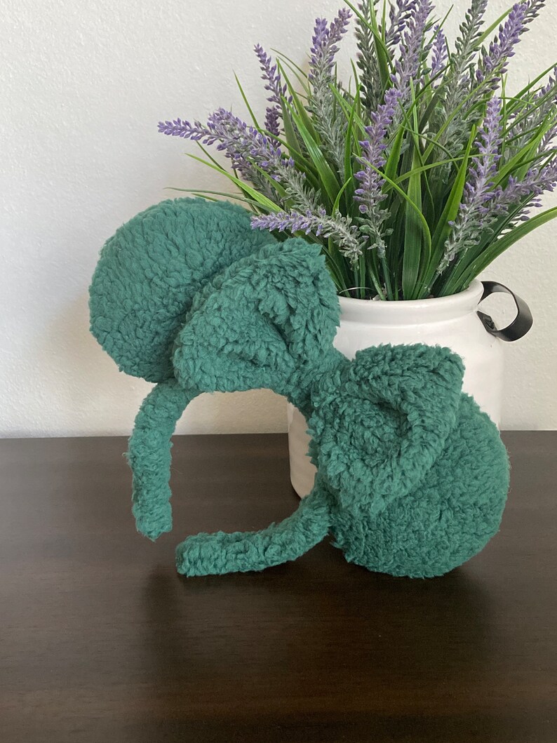 K&ouml;nnte beinhalten: T&uuml;rkisfarbenes Pl&uuml;sch-Mausohren-Stirnband. Die runden, flauschigen Ohren und das Stirnband sind t&uuml;rkisfarben. Das Stirnband liegt auf einer dunkelbraunen Oberfl&auml;che, mit einer wei&szlig;en Vase mit Lavendel im Hintergrund.