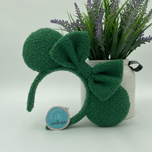K&ouml;nnte beinhalten: Ein gr&uuml;nes, flauschiges Stirnband mit einer gro&szlig;en Schleife und M&auml;useohren. Das Stirnband hat ein Etikett mit der Aufschrift "Handmade Ah Antikapr".