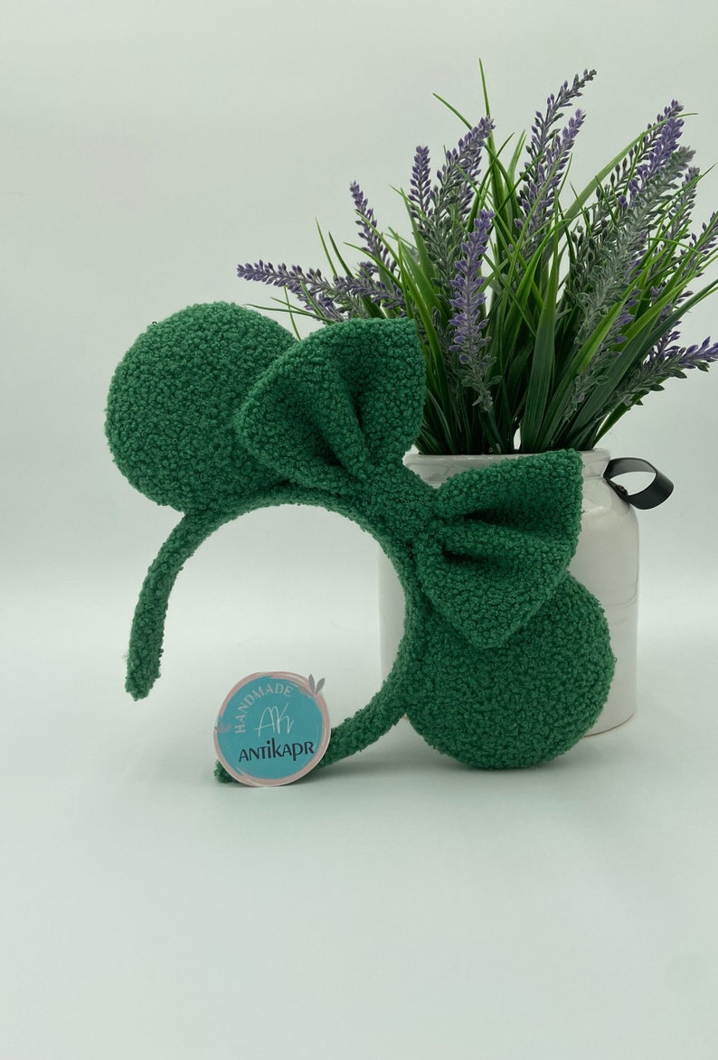 K&ouml;nnte beinhalten: Ein gr&uuml;nes, flauschiges Stirnband mit einer gro&szlig;en Schleife und M&auml;useohren. Das Stirnband hat ein Etikett mit der Aufschrift "Handmade Ah Antikapr".