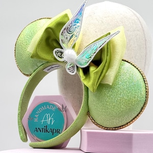 May include: Green and gold headband with large, glittery ears. A large green bow and iridescent butterfly wings are centered on the headband. The ears are outlined with gold trim. The headband is displayed on a mannequin head.