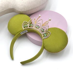 May include: A headband featuring two round, olive green velvet ears and a central gold-tone tiara. The tiara is embellished with clear crystals, small green marquise-cut stones, and white pearl accents. A small white tag reads ANTIKAPR.