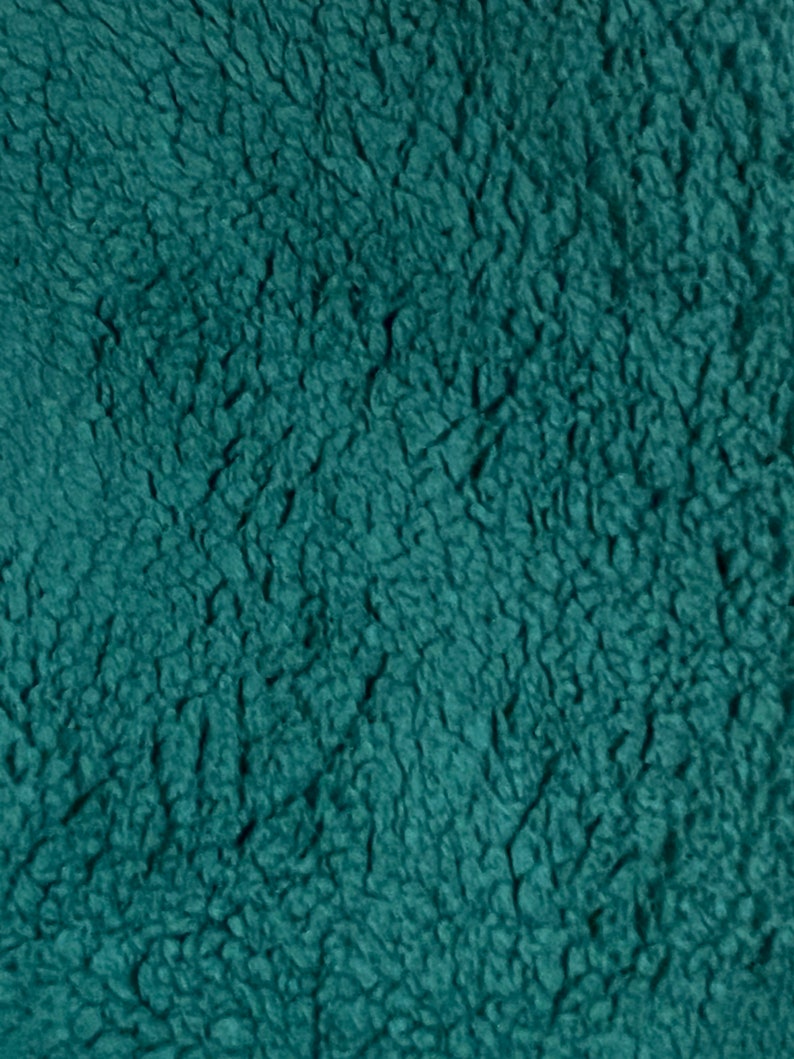K&ouml;nnte beinhalten: Nahaufnahme eines teal-farbenen, strukturierten Stoffes. Das Material hat ein pl&uuml;schiges, zotteliges Aussehen mit einer dichten, unebenen Oberfl&auml;che. Die Textur des Stoffes erinnert an ein weiches, warmes Material, m&ouml;glicherweise eine Decke oder ein Kleidungsst&uuml;ck.
