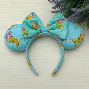May include: Light blue Minnie Mouse ears with a large bow featuring a print of the Seven Dwarfs from Snow White and the Seven Dwarfs. The ears are covered in a light blue glitter.