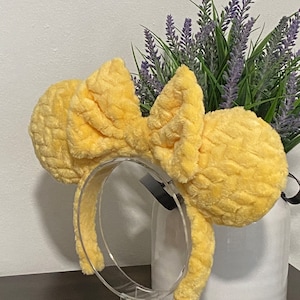 May include: Yellow fuzzy Minnie Mouse ears headband with a large bow. The headband is on a clear plastic stand.
