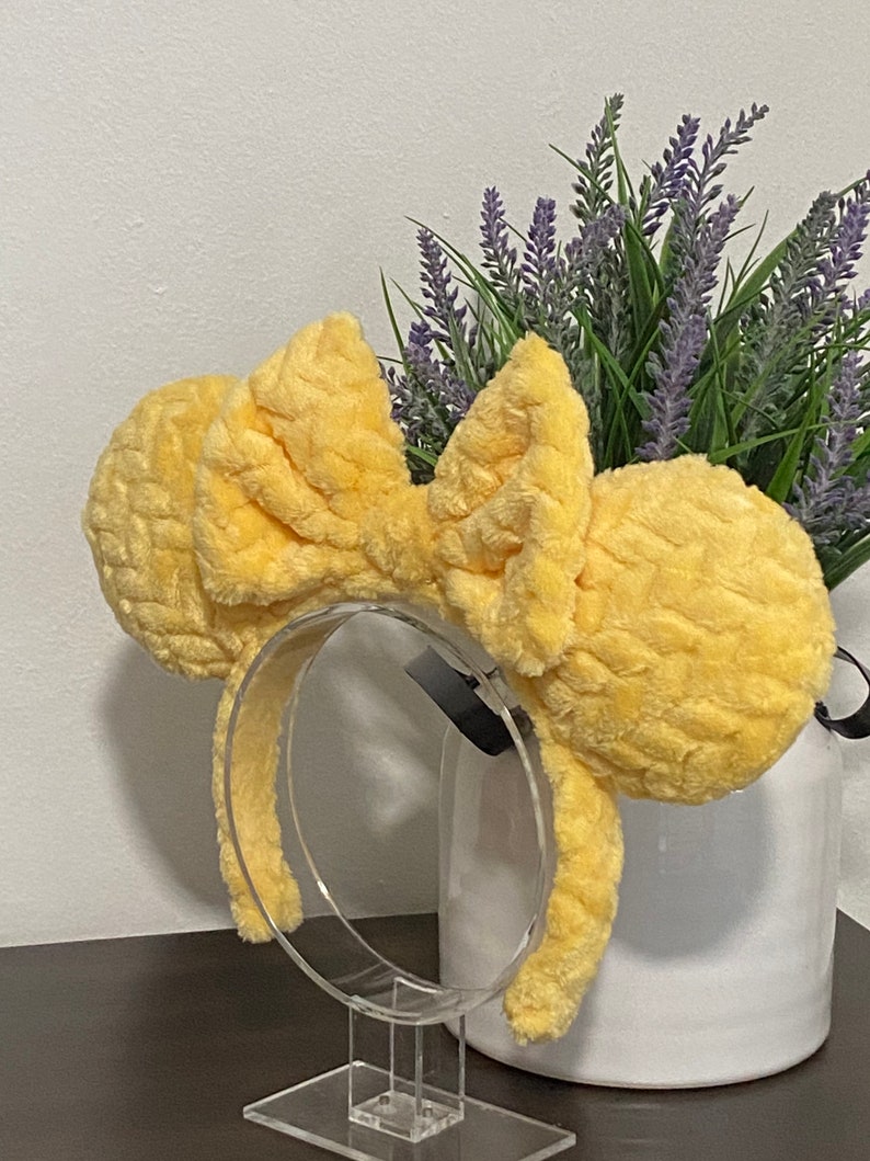 May include: Yellow fuzzy Minnie Mouse ears headband with a large bow. The headband is on a clear plastic stand.