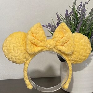 May include: Yellow fuzzy Minnie Mouse ears with a large bow. The ears are on a clear plastic stand.