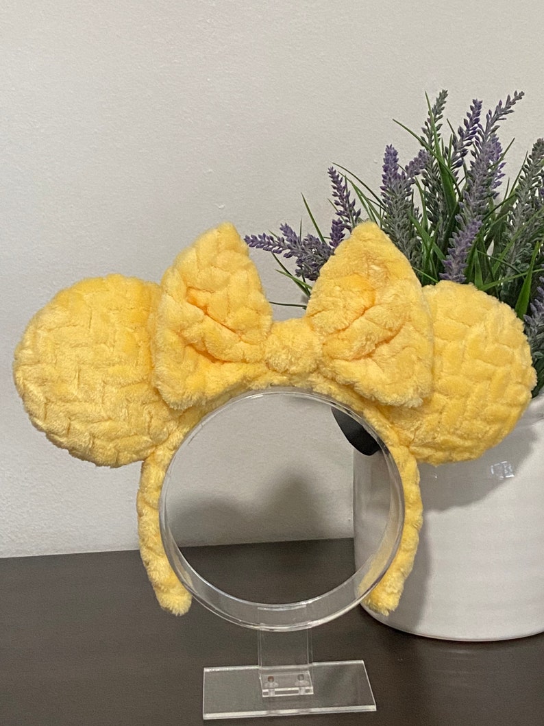 May include: Yellow fuzzy Minnie Mouse ears with a large bow. The ears are on a clear plastic stand.