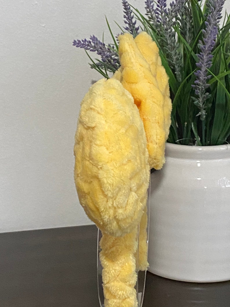 May include: Two yellow plush headbands with a fuzzy texture. The headbands are on a clear display stand.