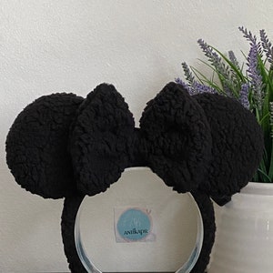 May include: Black fuzzy Minnie Mouse ears headband with a large black bow. The headband is on a clear stand and the background is white.
