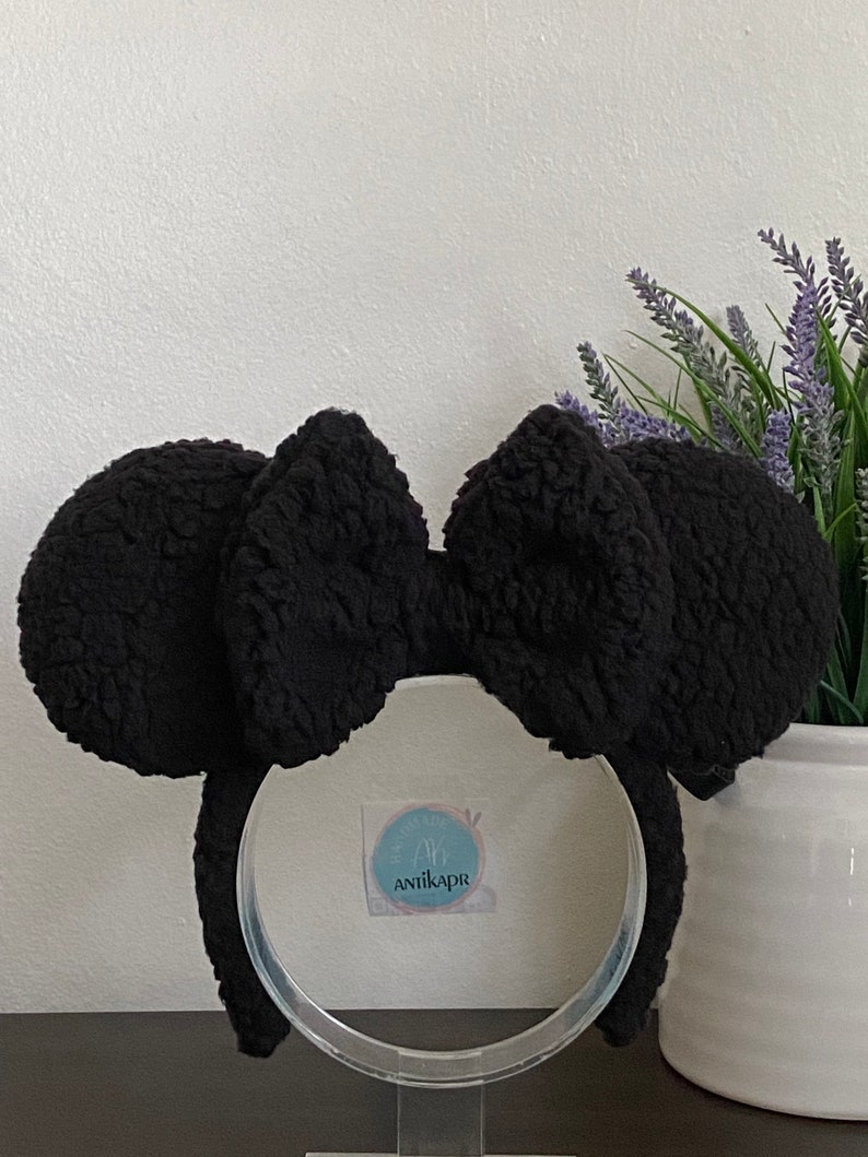 May include: Black fuzzy Minnie Mouse ears headband with a large black bow. The headband is on a clear stand and the background is white.
