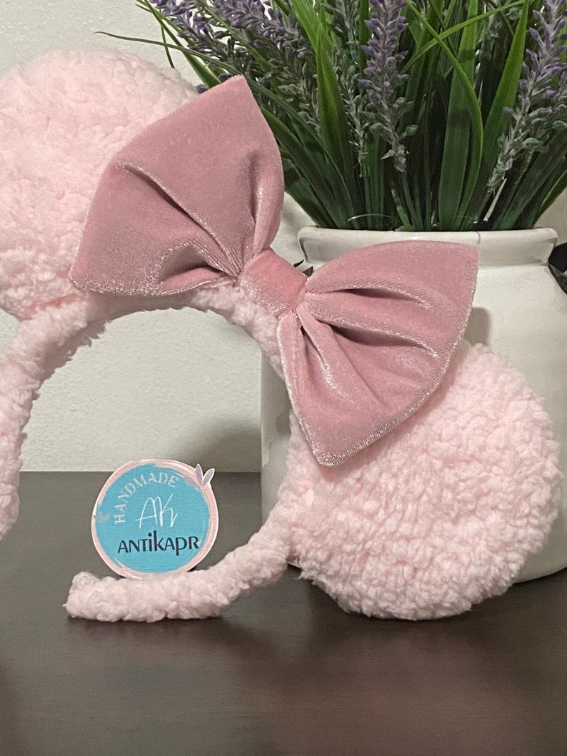 May include: Pink fuzzy mouse ears headband with a large pink velvet bow. The headband has a small blue circle tag with the text "Handmade Ah Antikapr".