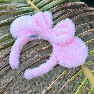 Op de afbeelding: Een roze pluche hoofdband met konijnenoren. De hoofdband heeft een wit label met het woord "ANTIKAPR" erop.