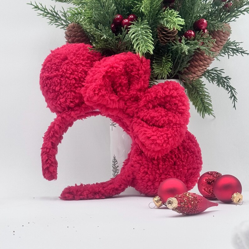 Puede incluir: Una diadema roja brillante y esponjosa con grandes orejeras en forma de oreja y un lazo. La diadema se exhibe con adornos navide&ntilde;os y vegetaci&oacute;n, creando un tema festivo de vacaciones.