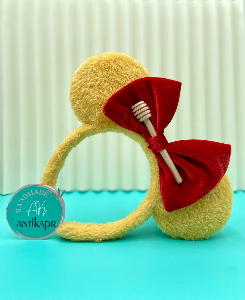 Pu&ograve; includere: Una fascia per capelli gialla e morbida con un grande fiocco di velluto rosso e un accento di cucchiaio di miele in legno. La fascia ha due forme di orecchie rotonde. Il logo "HANDMADE AK ANTIKAPR" &egrave; su un cerchio turchese.