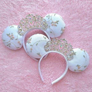 May include: Two white headbands featuring round ears with gold and green floral embroidery. Each headband is adorned with a large, intricate silver-tone tiara encrusted with clear, iridescent crystals and a central heart-shaped gem.