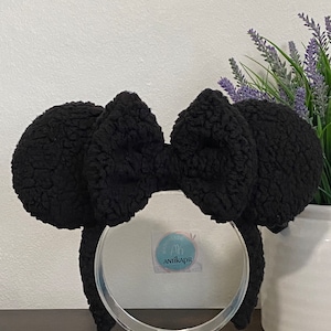 May include: Black fuzzy Minnie Mouse ears headband with a large black bow. The headband is on a clear plastic stand.