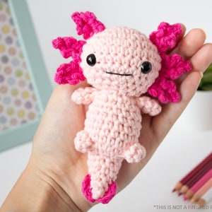 May include: A pink crocheted axolotl amigurumi toy with pink gills and black eyes. The toy is being held in a person's hand.