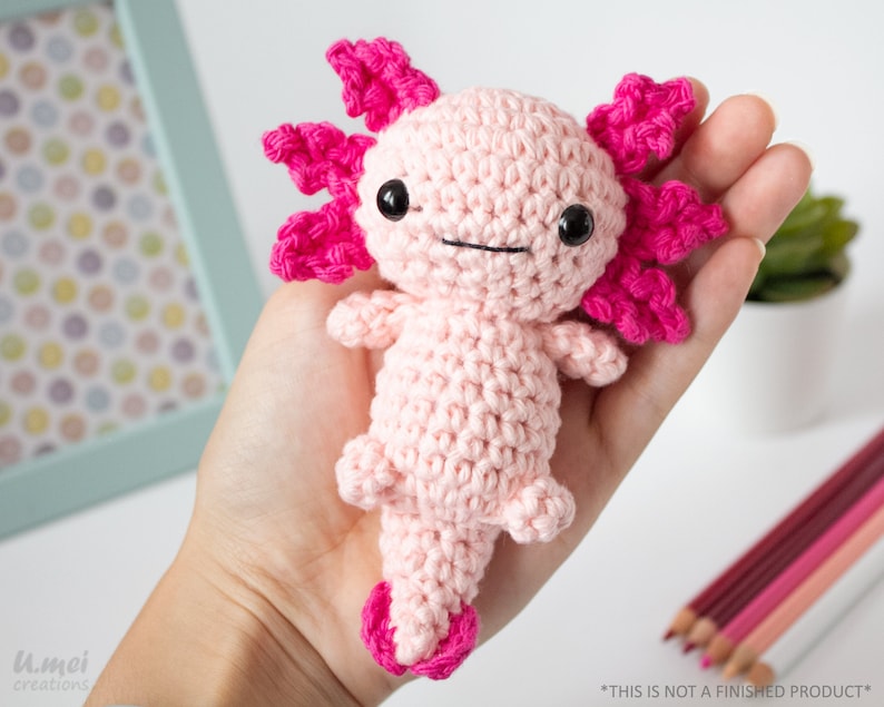 May include: A pink crocheted axolotl amigurumi toy with pink gills and black eyes. The toy is being held in a person's hand.