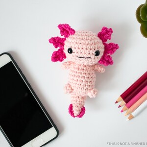 May include: Pink crocheted axolotl amigurumi toy with black eyes and a pink tail. The toy is on a white surface with a mobile phone and coloured pencils.