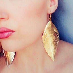 May include: A pair of gold feather earrings. The earrings are long and dangle from the earlobes. The feathers are made of a shiny, metallic material.
