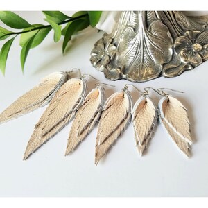 May include: Six pairs of beige leather feather earrings with silver-colored accents. The earrings are arranged in a row, showcasing their layered design and intricate details. The earrings are on a white surface.