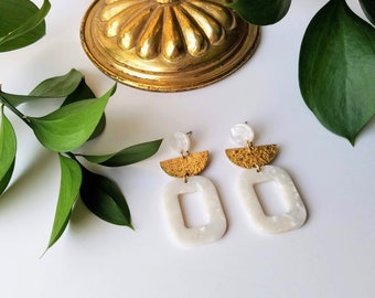 May include: A pair of gold and white geometric earrings. The earrings feature a large white oval shape with a smaller oval cutout in the centre. The top of the earrings feature a gold crescent shape with a textured surface.