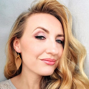 May include: A woman with long, wavy, light blonde hair is wearing a pair of brown leather earrings. The earrings are shaped like a leaf and have a gold-colored hook.