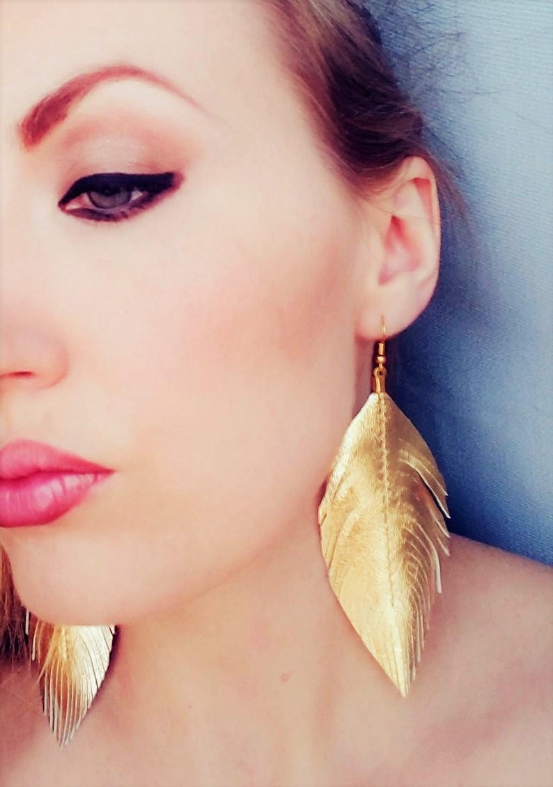 May include: A pair of gold feather earrings. The earrings are long and dangle from the earlobes. The feathers are made of a shiny, metallic material.