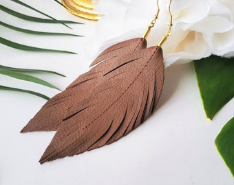 May include: A pair of brown leather feather earrings with gold-toned hooks. The earrings are shown against a white background with green leaves.