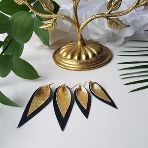 May include: A pair of gold and black leather earrings with a teardrop shape. The earrings are hanging from a gold earring hook.