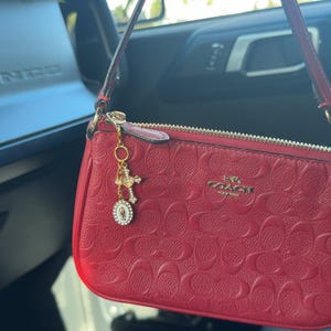May include: A red Coach wristlet with a gold zip and a gold charm. The wristlet features a repeating pattern of the Coach logo. The charm includes a cross and a religious medallion.